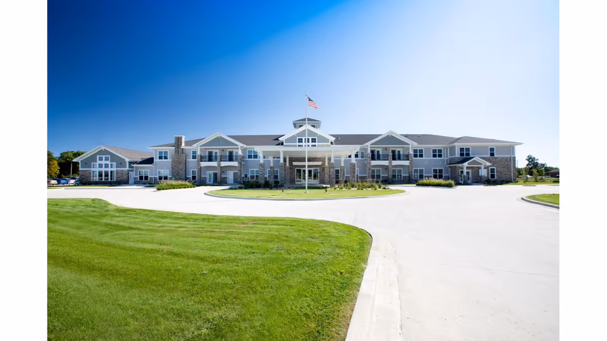 Front exterior of a two-story senior living building with a circular driveway, American flag, and green lawn under a clear blue sky.