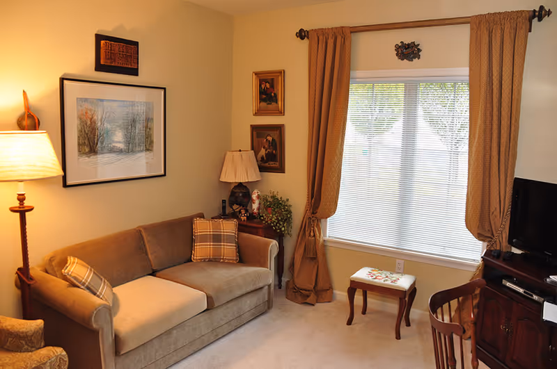 A cozy living room with a brown sofa adorned with plaid pillows, a floor lamp, framed artwork on the walls, a small wooden side table with a lamp and decorative items, a window with beige curtains and blinds, a small cushioned stool, a wooden chair, and a TV on a wooden stand.