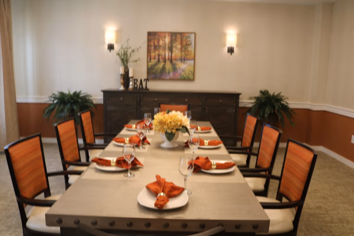 A dining room with a long rectangular table set for eight people. Each place setting has a white plate with an orange napkin and a wine glass. The chairs have orange-striped upholstery. A centerpiece with yellow flowers is in the middle of the table. In the background, there is a dark wooden sideboard with decorative items and a framed painting of a forest scene on the wall. Two wall sconces provide lighting, and there are green plants on either side of the sideboard.