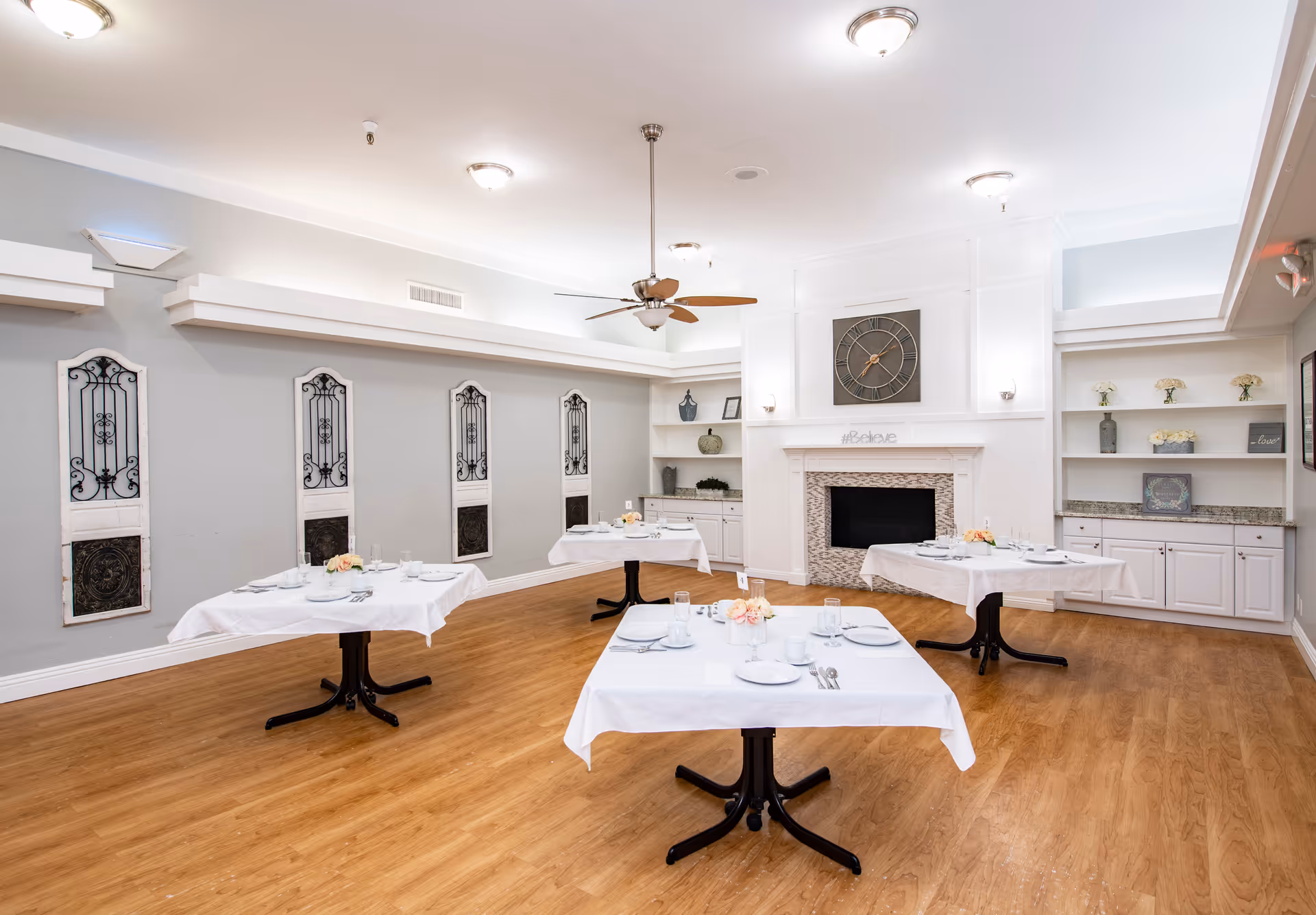 A bright dining room with four tables covered in white tablecloths, each set with plates, glasses, and cutlery. The room features wooden flooring, a ceiling fan, a large wall clock above a decorative fireplace, and built-in shelves with decorative items and flowers.