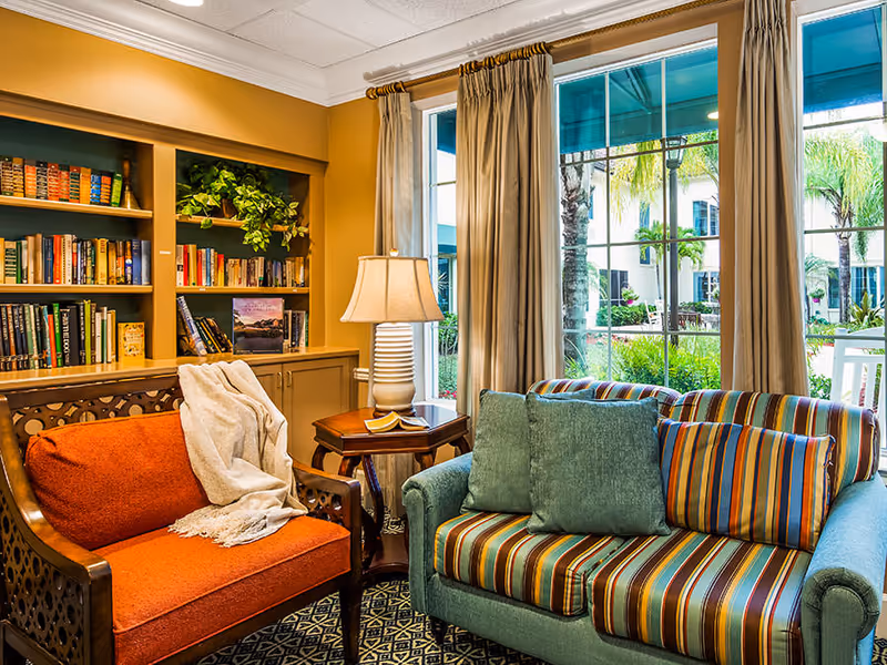 Cozy living room area with bookshelves, an orange armchair draped with a blanket, a striped teal loveseat, a side table with a lamp, and large windows looking out to a courtyard.