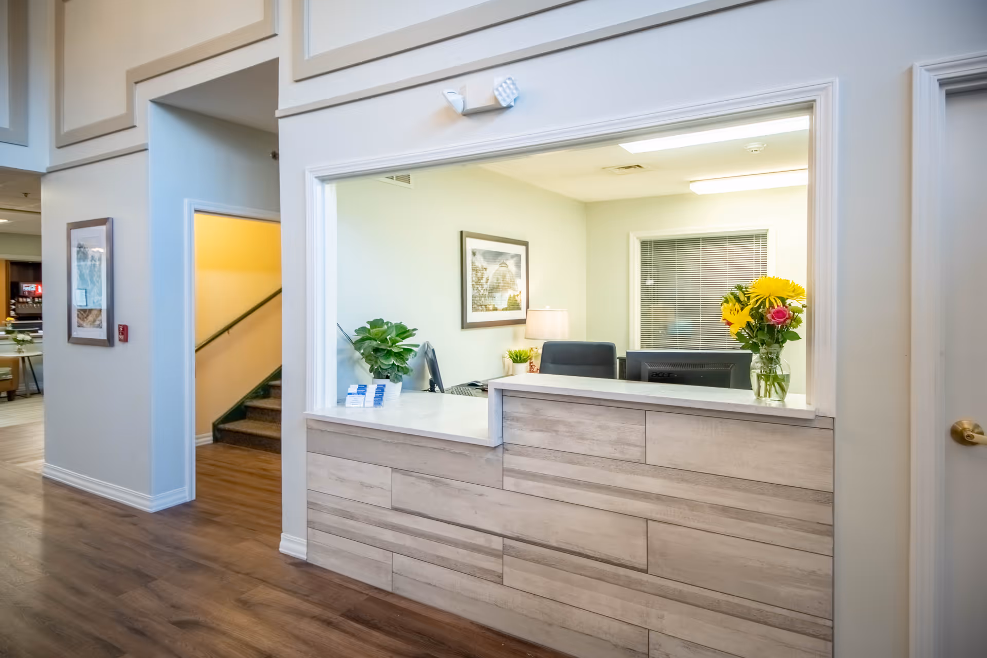 Reception area with a wooden front desk, a computer monitor, a vase with yellow and pink flowers, a potted plant, and a framed picture on the wall behind the desk. To the left, there is a hallway with stairs and a framed picture on the wall.