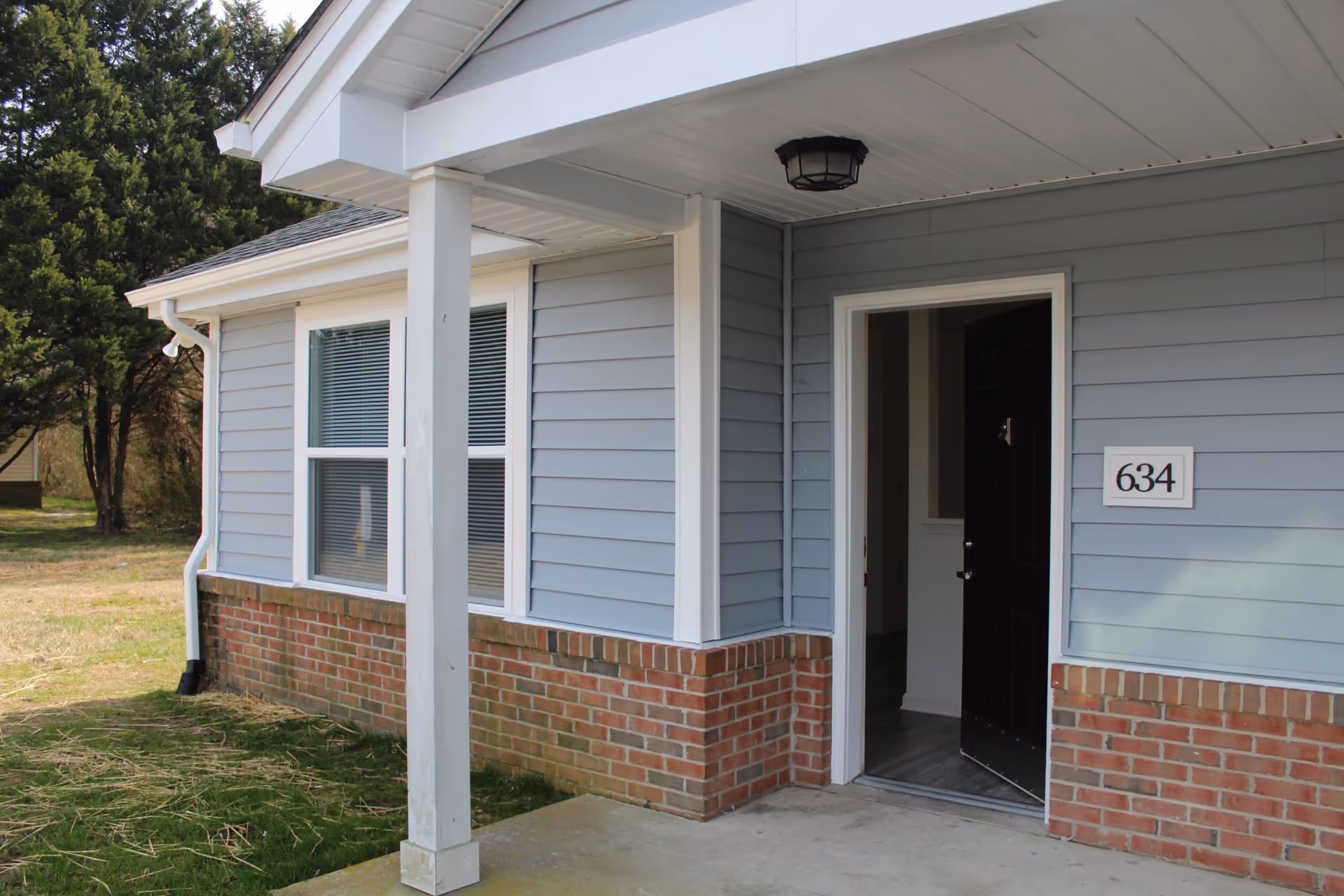 Exterior view of a residential building with light blue siding and red brick accents. The building has a covered porch supported by a white column and a black door that is partially open. The house number 634 is displayed next to the door. There are two windows with white frames and blinds on the left side. Trees and grass are visible in the background.