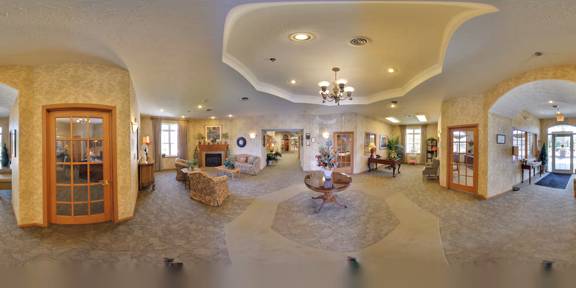 A spacious and warmly decorated senior living facility common area with beige patterned wallpaper and carpet. The room features a central round wooden table with a floral arrangement, multiple seating areas with sofas and armchairs, a fireplace with a mantel decorated with plants and artwork, and several wooden doors with glass panels. The ceiling has recessed lighting and a chandelier. There are windows letting in natural light and various decorative plants and framed pictures on the walls.
