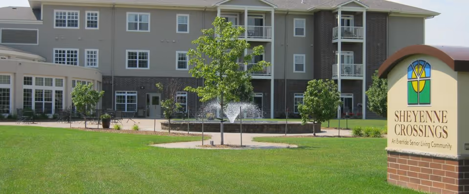 Exterior view of a senior living community building with multiple windows and balconies. In front of the building, there is a green lawn with young trees and a circular water fountain. A sign on the right side reads 'Sheyenne Crossings, An Eventide Senior Living Community.'