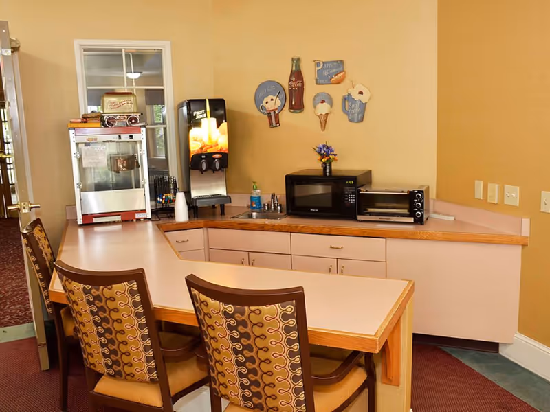 A small kitchenette area with a counter and three patterned chairs. On the counter are a popcorn machine, a beverage dispenser, a microwave, a toaster oven, and a small sink. The wall behind has decorative signs featuring ice cream, a Coca-Cola bottle, and a hot dog.