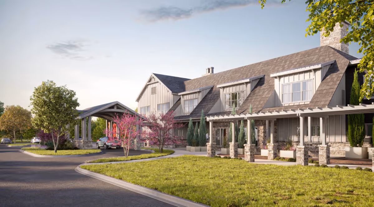 Exterior view of a senior living facility building with a covered entrance driveway, landscaped greenery including trees and bushes, and a clear sky in the background.