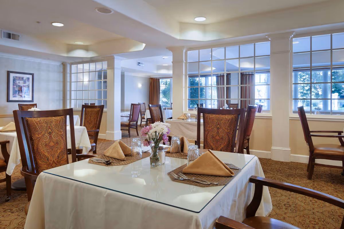 A well-lit dining room with tables covered in white tablecloths and glass tops, each set with folded beige napkins, glasses, and silverware. The chairs have wooden frames with patterned upholstery. There are large windows with white frames allowing natural light to enter, and a vase with flowers is placed on the table in the foreground.