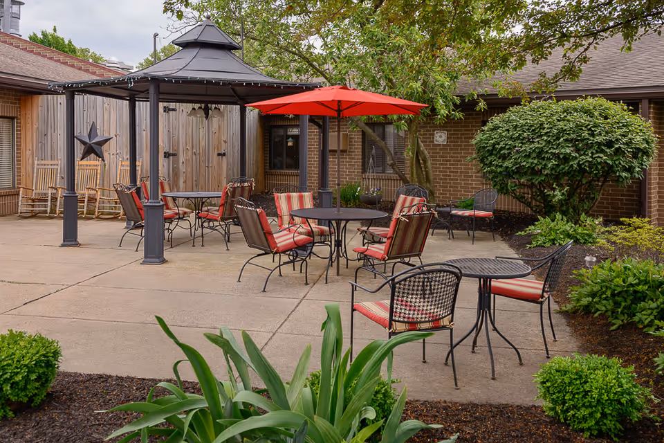 Outdoor patio area with several metal tables and chairs featuring red and striped cushions. A black gazebo and a red umbrella provide shade. Surrounding the patio are green bushes, plants, and trees, with a brick building and wooden fence in the background.