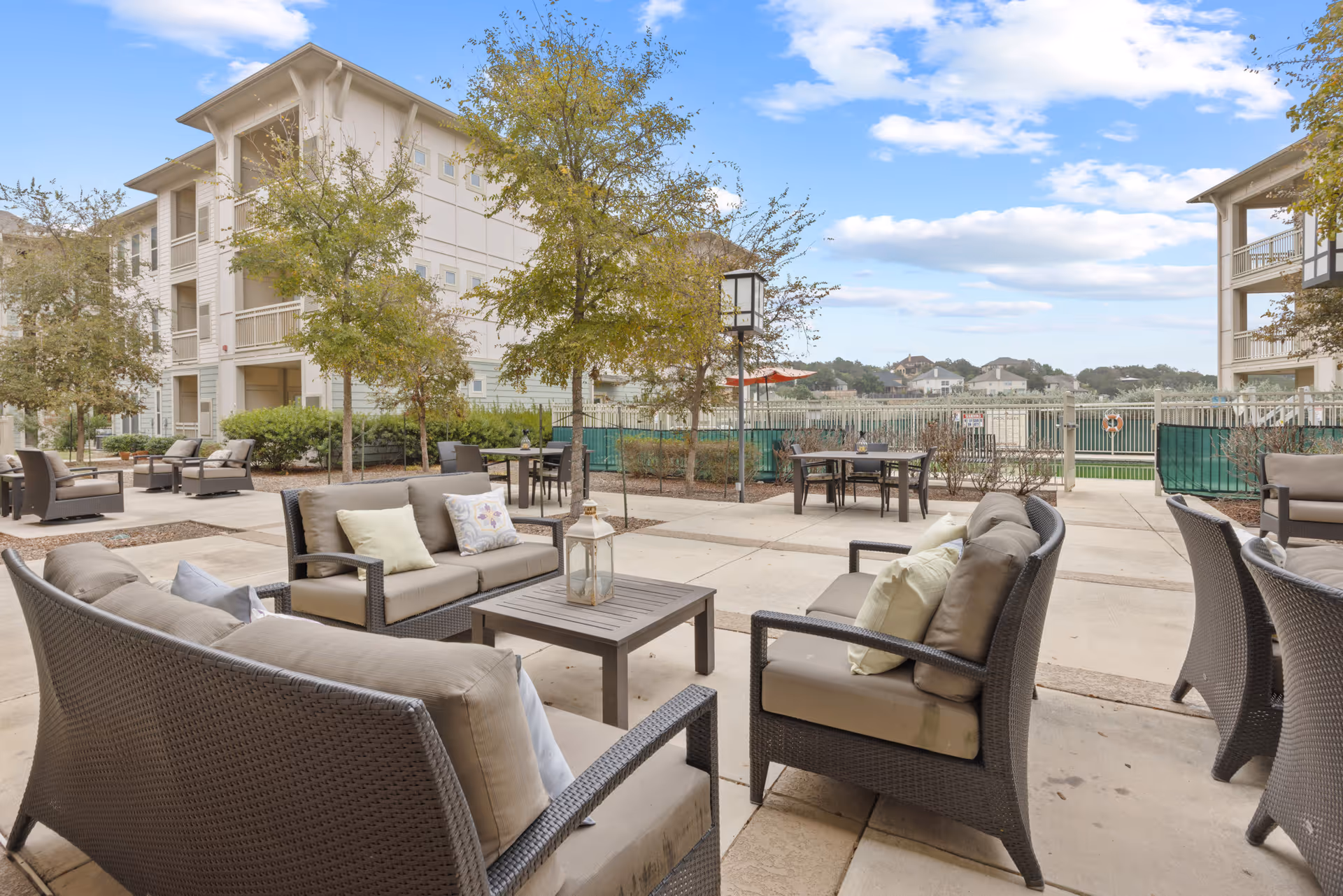 Outdoor patio area at Discovery Village Stone Oak with multiple cushioned wicker sofas and chairs arranged around low tables. Trees and shrubs are planted around the patio, and multi-story residential buildings are visible in the background under a partly cloudy sky.