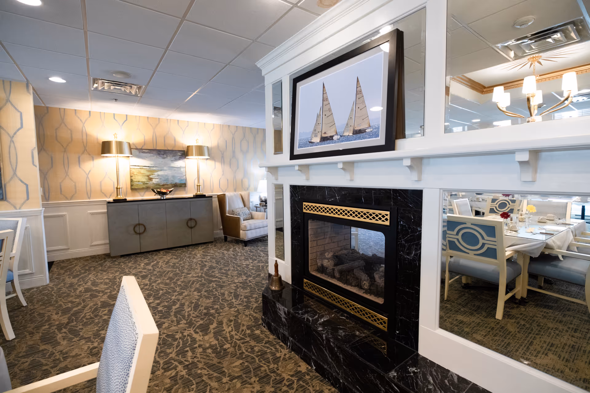 Interior view of a senior living facility dining area with a black marble fireplace in the foreground topped with a framed picture of two sailboats. The room features patterned carpet, wallpaper with a geometric design, a sideboard with two lamps, an armchair, and a dining table with chairs reflected in a mirror behind the fireplace.