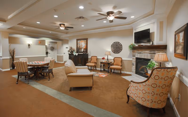 A cozy senior living facility common area with beige walls and carpeted floor. The room features a seating arrangement including a leather sofa, patterned armchairs, and wooden chairs around a small table with a checkerboard game. There is a stone fireplace with a TV mounted above it, decorative wall art, and table lamps providing warm lighting.