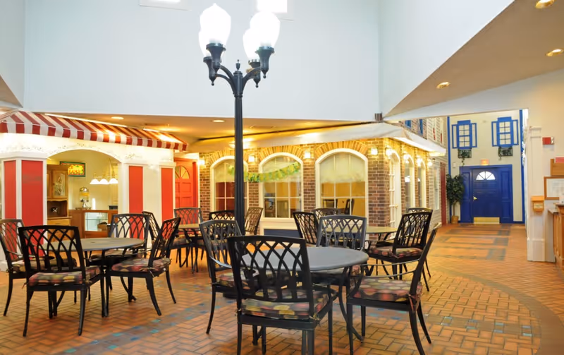 Indoor seating area with multiple round tables and black metal chairs with patterned cushions. The space features a streetlamp-style light fixture in the center, brick flooring, and walls designed to look like storefronts with windows and doors. The area has a bright and welcoming atmosphere with a high ceiling and recessed lighting.