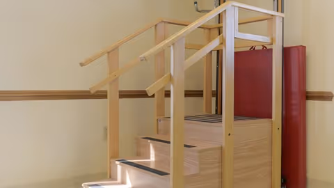 A wooden physical therapy staircase with handrails on both sides and non-slip strips on each step, placed indoors against a beige wall with a wooden chair rail. A red padded mat is positioned vertically behind the staircase.