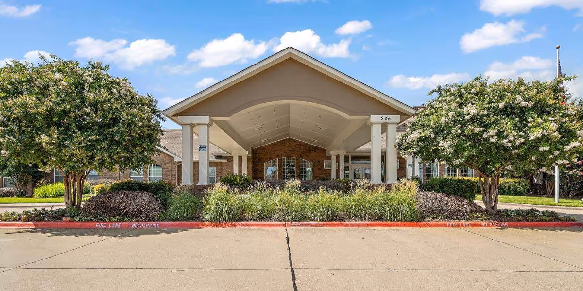 Front exterior view of Morada Cedar Hill facility showing a covered entrance with white pillars, surrounded by landscaped greenery and trees under a blue sky with scattered clouds.