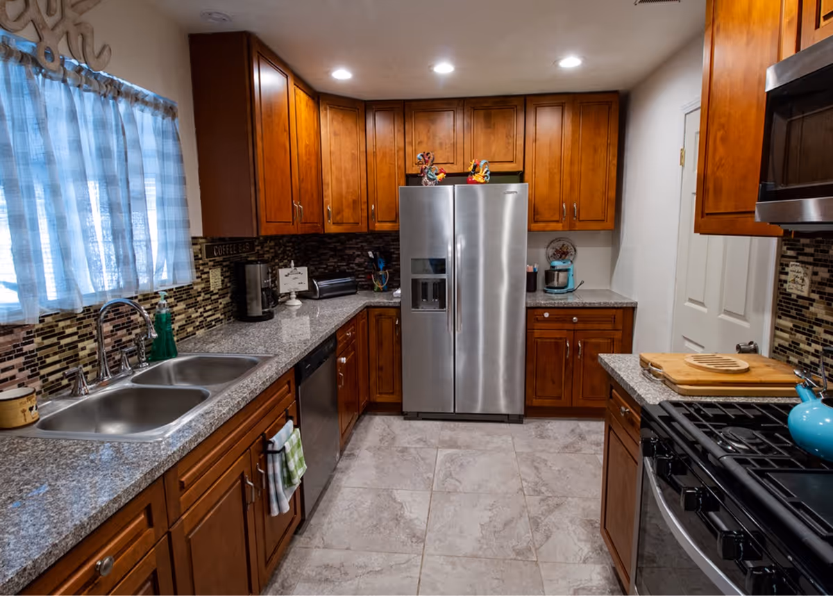 A modern kitchen with wooden cabinets, granite countertops, a double sink under a window with blue checkered curtains, a stainless steel refrigerator, dishwasher, and stove. The backsplash features a mosaic tile design, and various kitchen items like a coffee maker, toaster, and cutting board are visible on the counters.