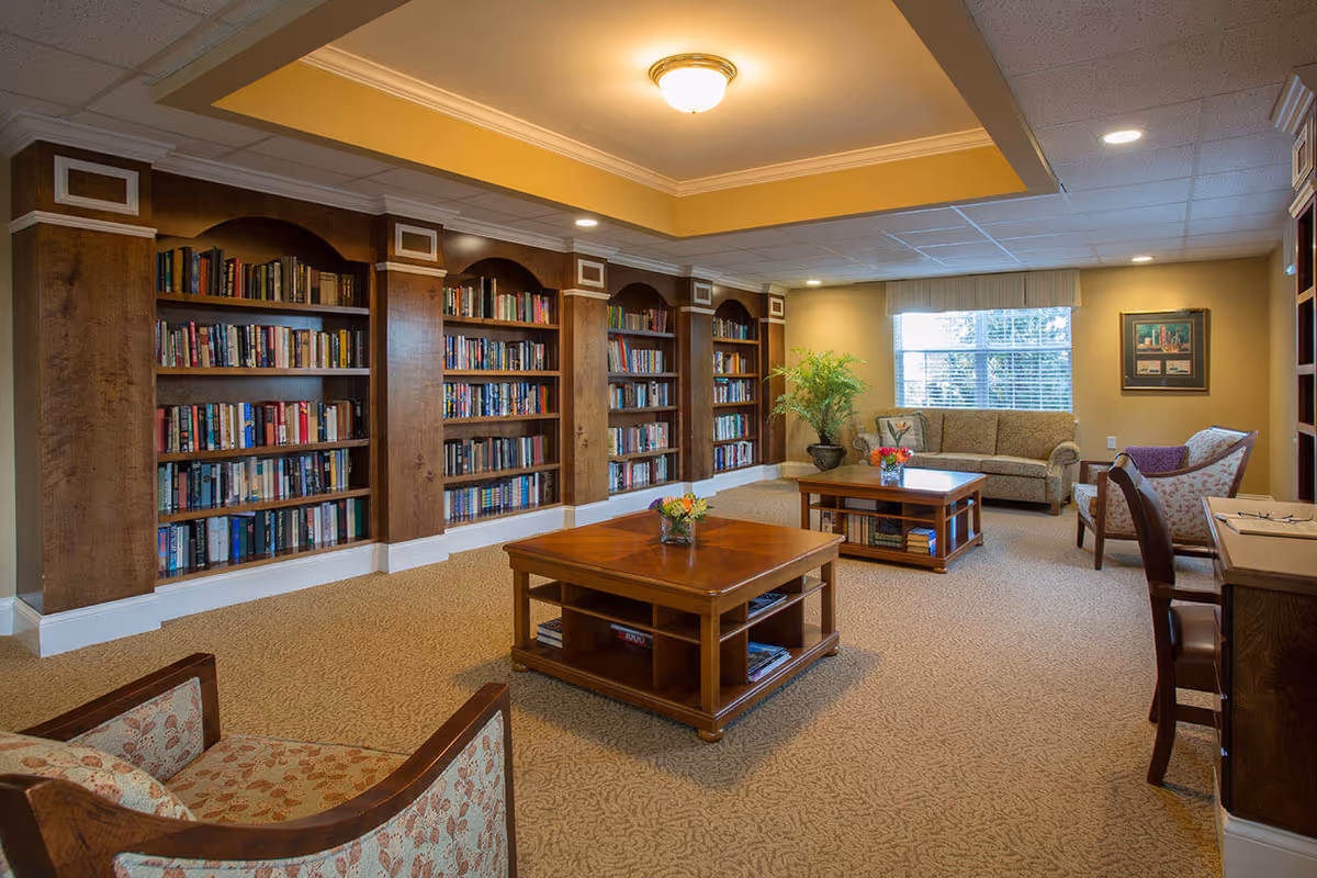 A cozy library or reading room with built-in wooden bookshelves filled with books along one wall, two wooden coffee tables with flowers on top, a sofa and armchairs arranged around the tables, a window with blinds letting in natural light, and a potted plant in the corner.