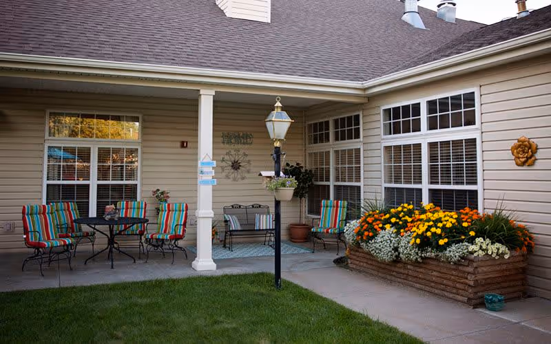 Outdoor patio area at Bickford of Grand Island featuring colorful striped chairs around a black metal table, a bench with cushions, a lamp post with a hanging flower pot, and a large wooden planter box filled with vibrant yellow, orange, and white flowers. The patio is adjacent to a beige building with large windows and decorative wall hangings.