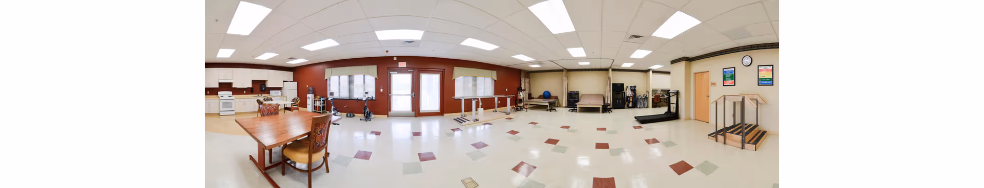 Wide interior view of a rehabilitation/activity room with tables and chairs, a kitchenette on the left, and exercise/therapy equipment including parallel bars.