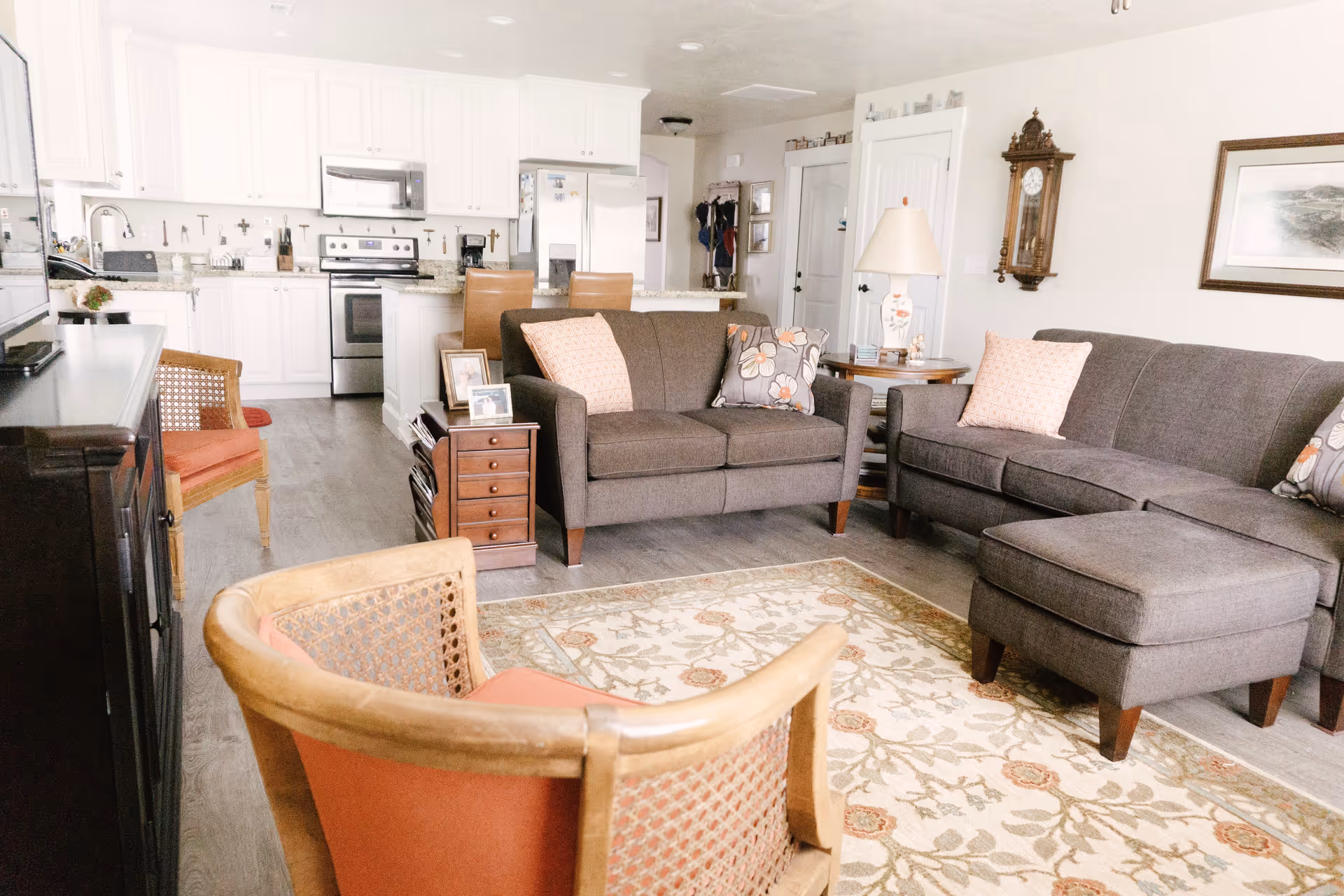 Open-plan living room with gray sofas, patterned rug, wooden chairs and a view into a white kitchen.