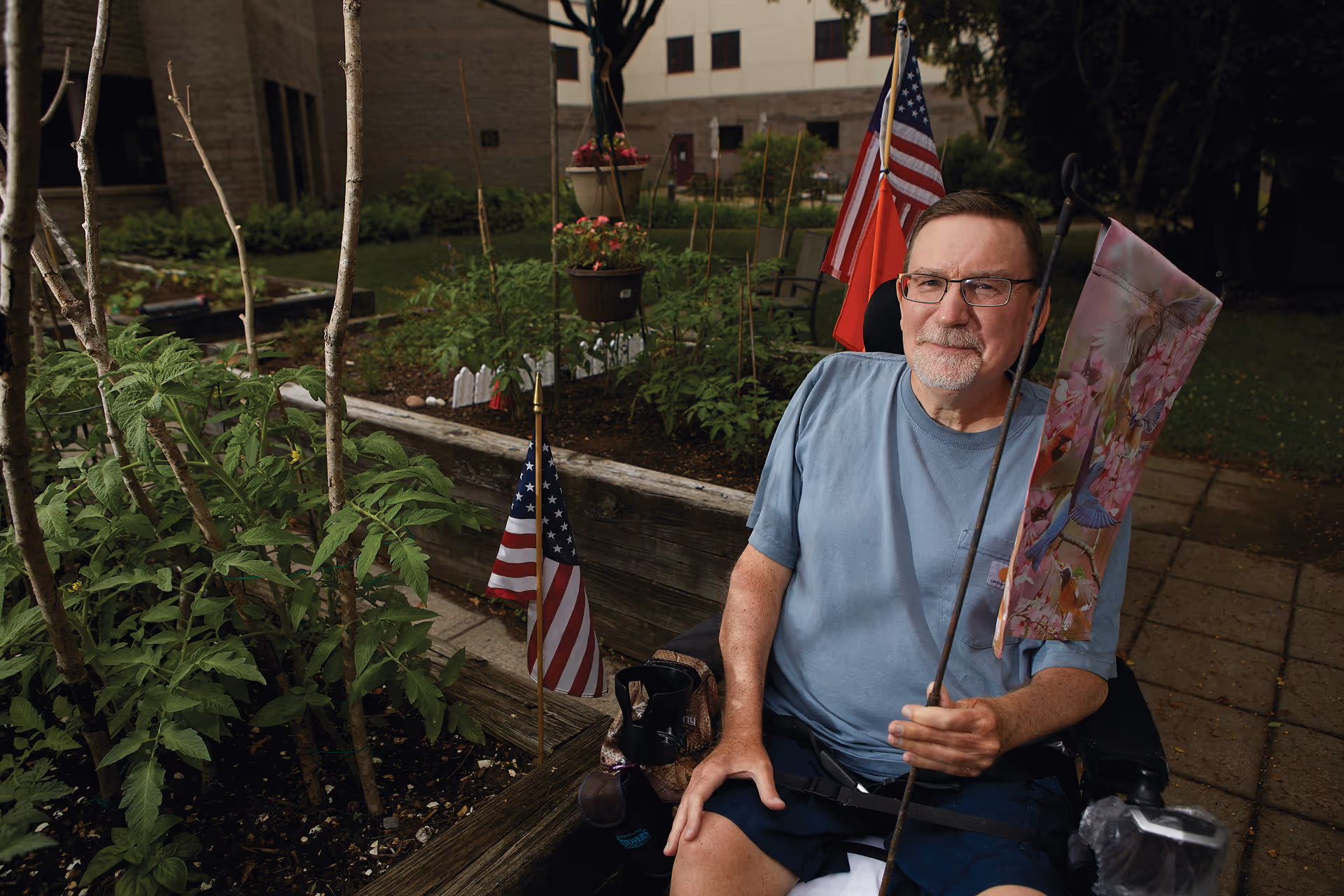 An elderly man in a wheelchair holding a decorative flag with flowers and birds, sitting next to a garden bed with plants and small American flags. The background shows a building and greenery.