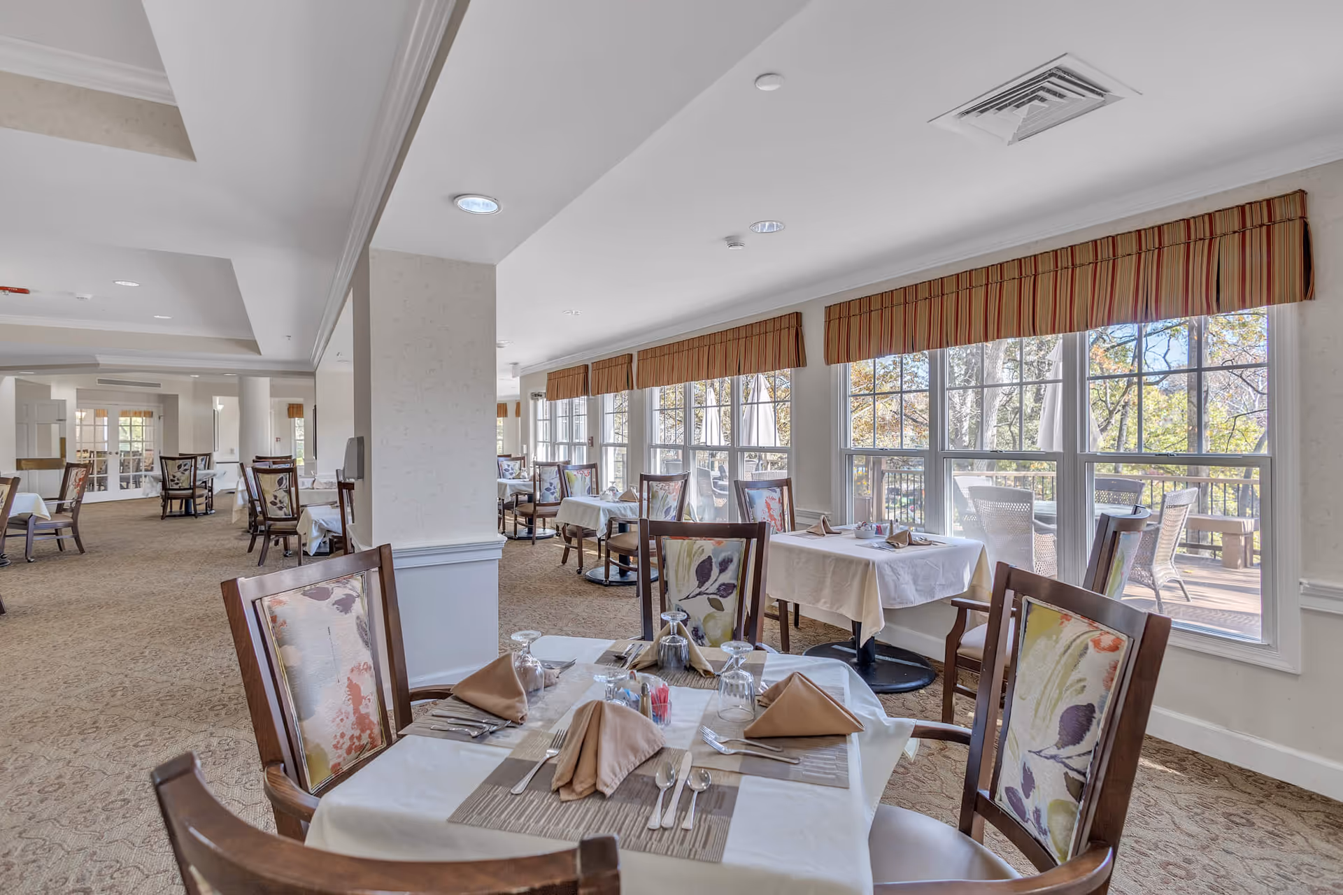 Bright dining room with tables set for service, upholstered chairs, and large windows overlooking an outdoor deck.