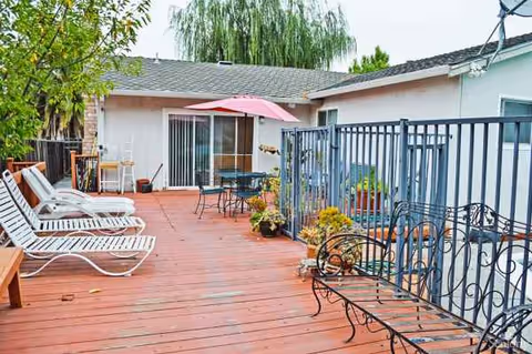 An outdoor senior living patio with lounge chairs, a table, umbrella, and potted plants.