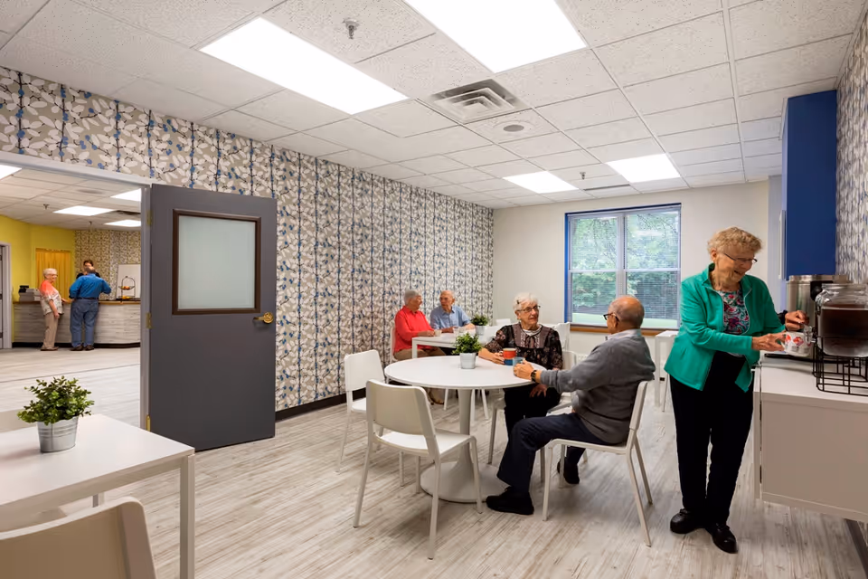 A bright common area in a senior living facility with several elderly people sitting at round white tables and chatting. One woman is standing near a counter pouring a drink from a dispenser. The room has patterned wallpaper, large windows, and light wood flooring. Another room with more people is visible through an open door.