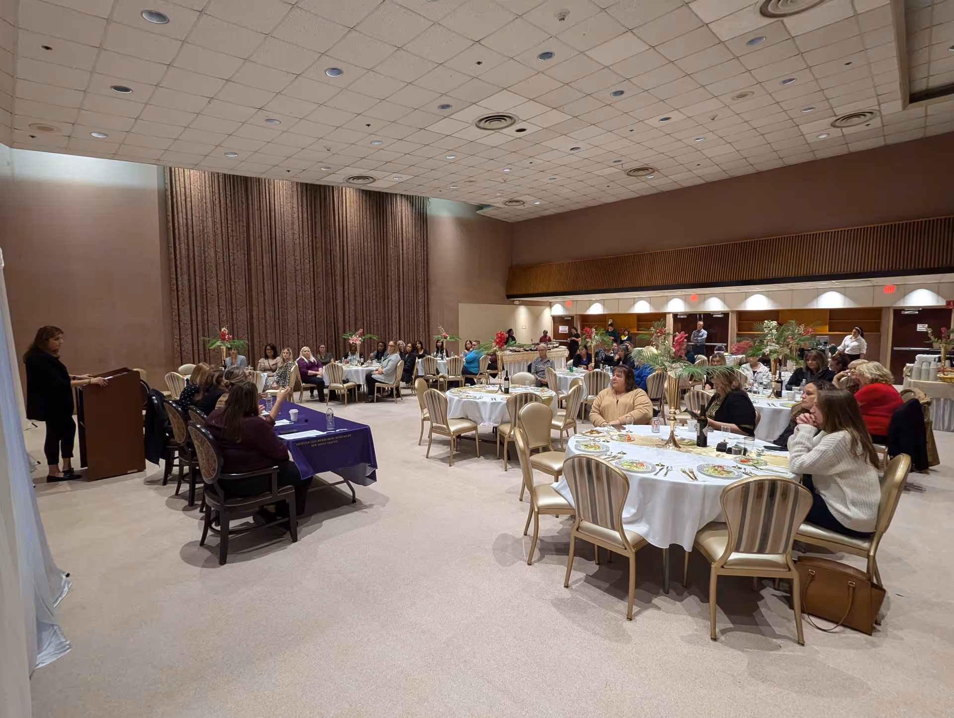 A large room set up for a formal event with round tables covered in white tablecloths and floral centerpieces. Several people are seated at the tables, listening to a woman speaking at a podium on the left side of the image. The room has a high ceiling with recessed lighting and beige walls with tall curtains.