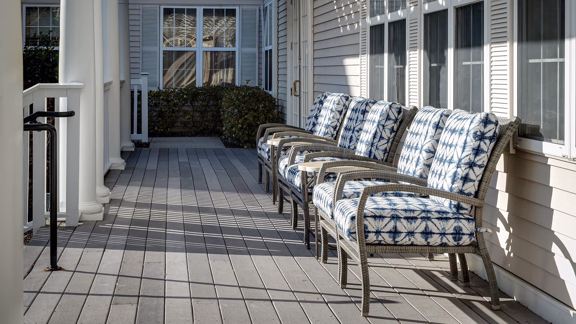 A sunlit outdoor porch area with a row of five cushioned wicker chairs featuring blue and white patterned cushions. The porch has white columns and railings, with windows and beige siding on the building wall beside the chairs.