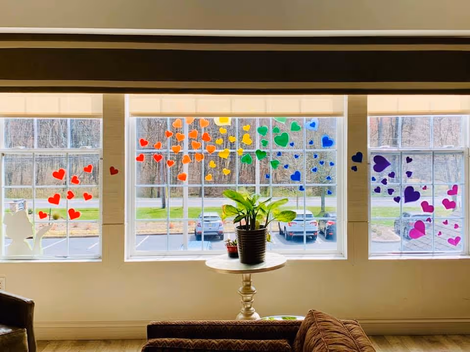 A bright room with three large windows decorated with colorful heart-shaped stickers arranged in a rainbow pattern. Outside the windows, a parking lot and trees are visible. Inside, there is a small round table with a potted plant and a smaller plant beside it. Part of a brown couch and a chair are also visible.