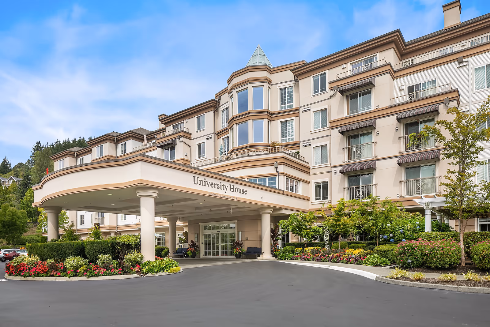 Front exterior view of University House Issaquah, a multi-story senior living facility with a covered entrance, landscaped gardens, and clear blue sky.