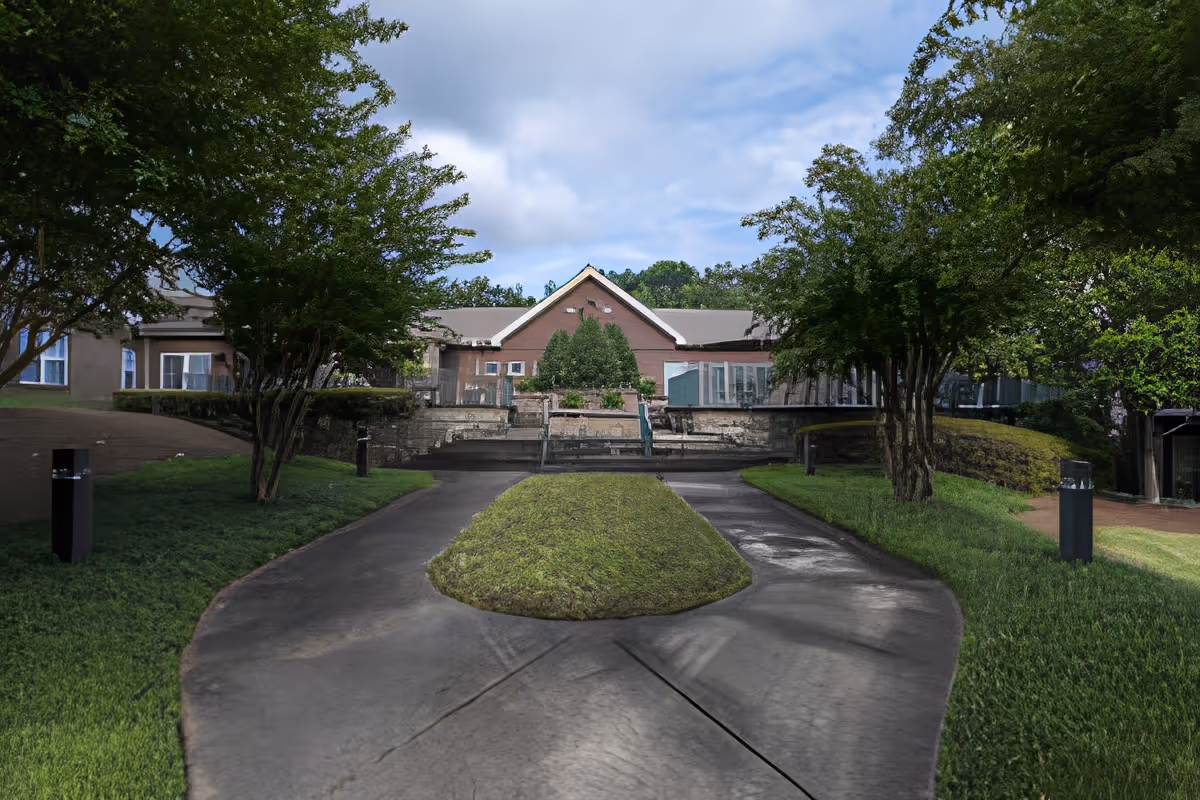 Paved walkway with a grassy center leading to the front of a red-brick building flanked by trees and landscaping.
