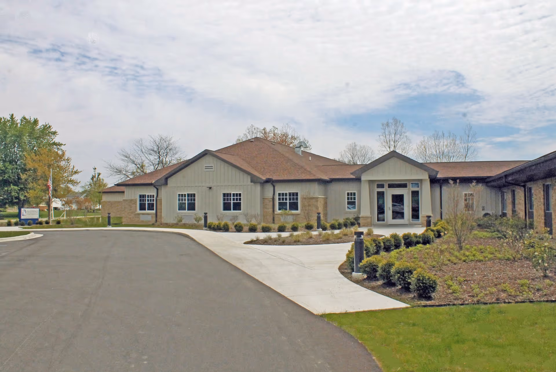 Exterior view of a single-story nursing and rehabilitation community building with a paved driveway, landscaped bushes, and a partly cloudy sky.