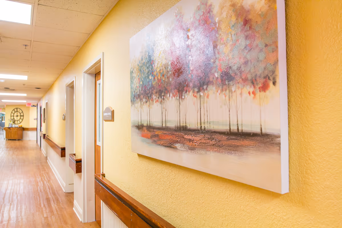 A brightly lit hallway with wooden flooring and yellow walls. A colorful painting of trees hangs on the right wall. Several doors line the hallway, one with a sign that reads 'Nurses Office'. At the end of the hallway, there is a large decorative clock on the wall above a wooden cabinet.