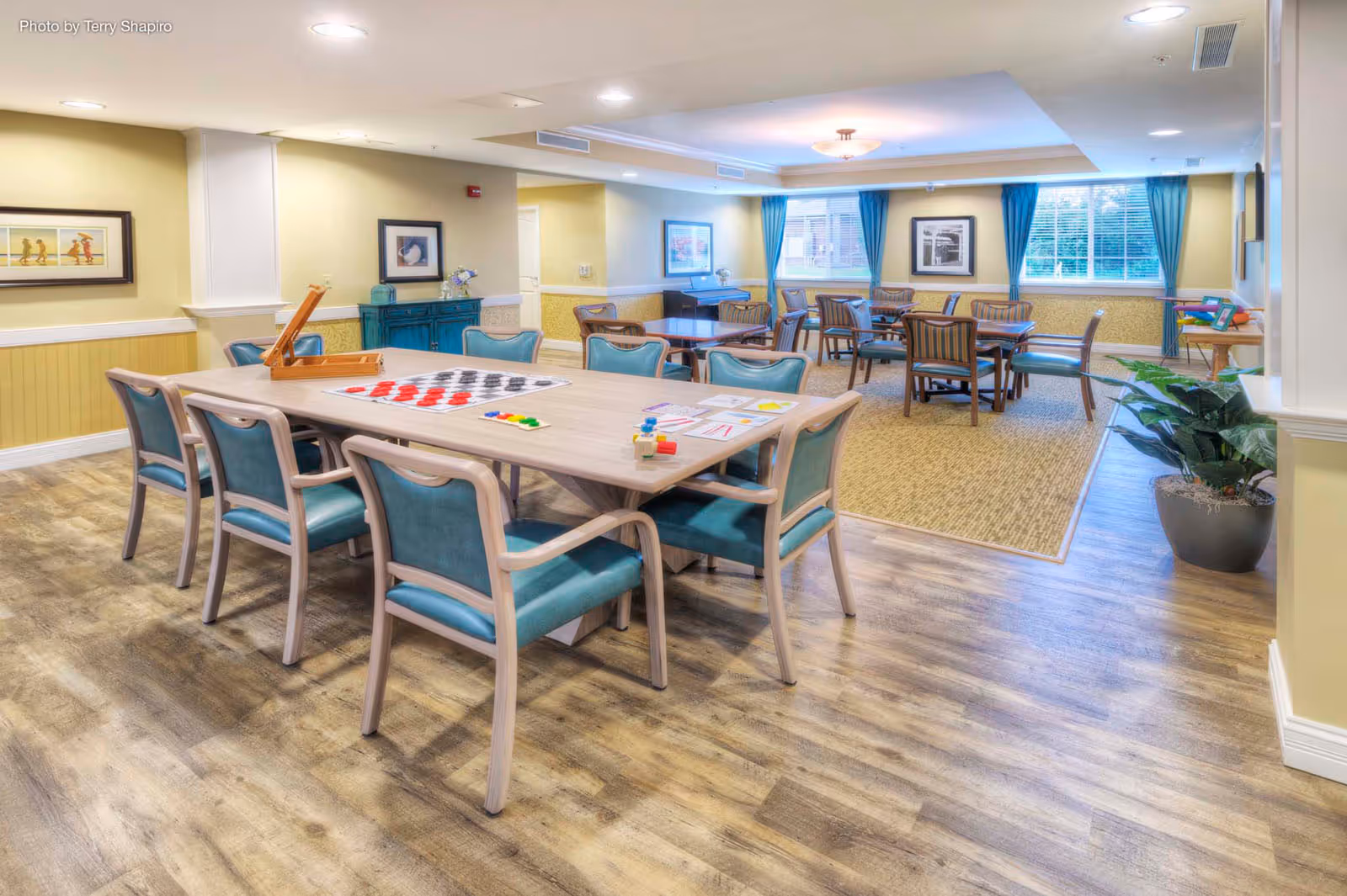 Bright communal activity/dining room with tables and teal-upholstered chairs and games set out on the central table.
