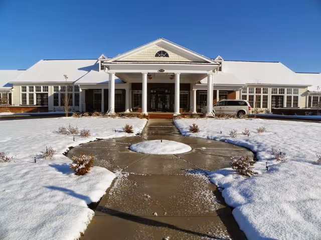 Snow-covered circular walkway leading to the columned front entrance of a low-rise building under a clear blue sky.