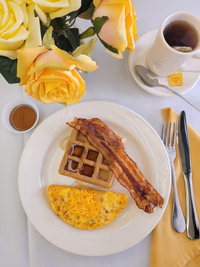 A breakfast plate with a folded omelette topped with shredded cheese, two strips of crispy bacon, and a waffle with syrup. Next to the plate is a cup of tea with a spoon on the saucer, a small container of syrup, and a yellow napkin with a fork and knife. Yellow roses in a vase are also visible on the table.