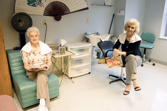 Two elderly women sitting in a hair salon area of a retirement residence. One woman is seated on a green chair holding a magazine, and the other is sitting on a salon chair holding a magazine. The room has a hair washing station, a fan decoration on the wall, and a small table with tissues and magazines.