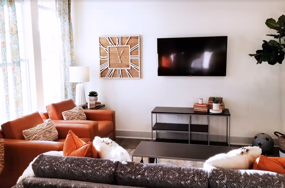 A cozy living room with a gray patterned sofa and two orange armchairs, each with decorative pillows. A large wall clock and a flat-screen TV are mounted on the white wall. A black console table beneath the TV holds stacked books and small plants. A floor lamp and a potted plant are also visible, with light streaming in through windows with floral curtains.