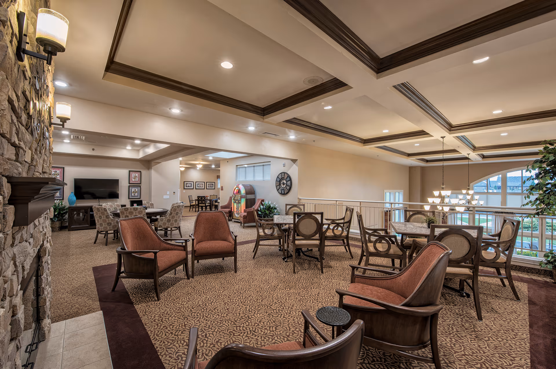 A spacious and well-lit common area in an assisted living facility featuring multiple seating arrangements with cushioned chairs and tables. The room has a patterned carpet, a stone fireplace on the left, a large wall clock, a jukebox, and a flat-screen TV mounted on the far wall. Large windows on the right side allow natural light to enter, and the ceiling has decorative wooden beams with recessed lighting.