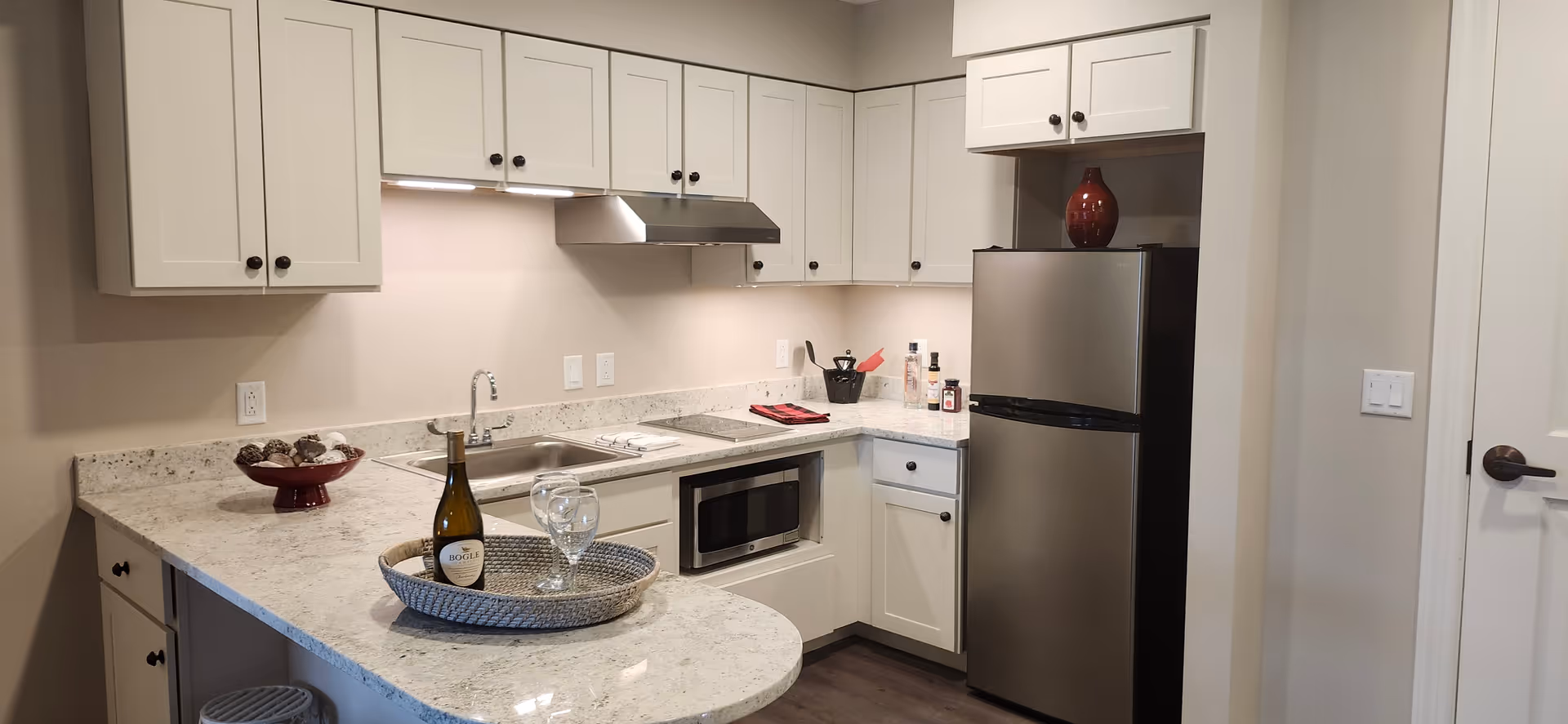 A modern kitchen with white cabinets, a stainless steel refrigerator, a microwave, and a sink. The countertop is light-colored granite with a bottle of wine and two wine glasses on a woven tray. There is a red decorative bowl with pine cones and kitchen utensils on the counter.