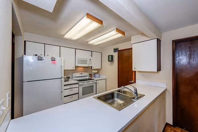 Small kitchen with white cabinets and appliances, a countertop peninsula with a stainless sink, and overhead fluorescent lights.
