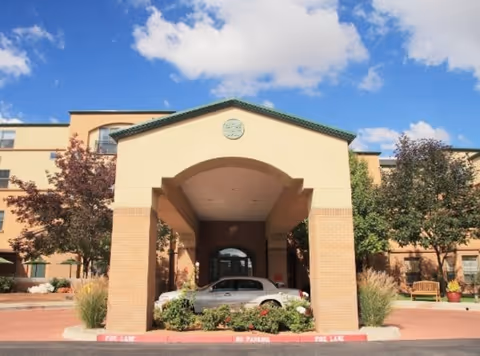 Entrance of a multi-story senior living facility with a covered driveway supported by brick columns. There are trees and landscaping around the entrance, a silver car parked under the covered area, and a clear blue sky with some clouds above.