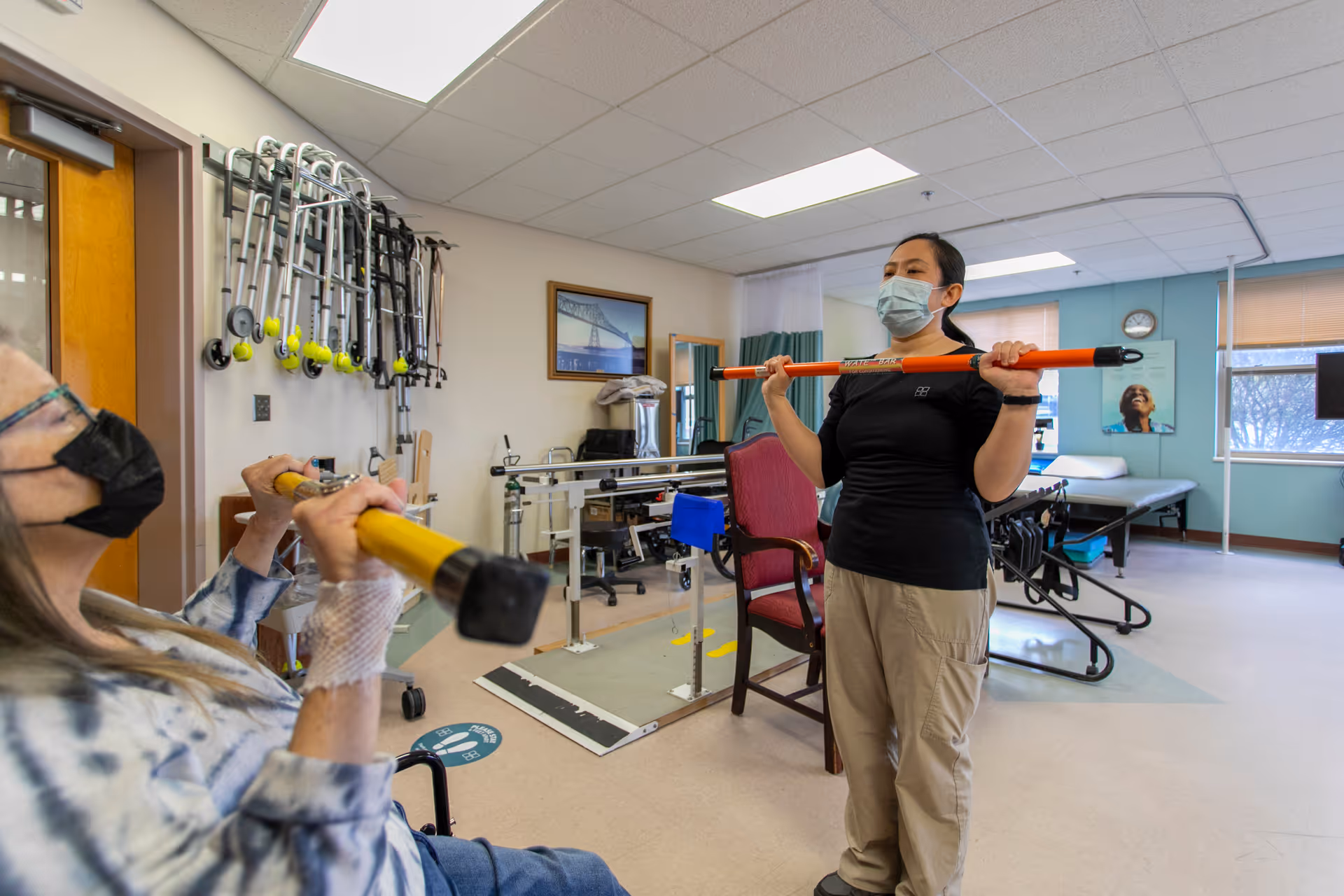 A physical therapy session in a rehabilitation room where a therapist wearing a mask is guiding an elderly woman, also masked, who is seated and holding a yellow exercise bar. The room contains various mobility aids like walkers hanging on the wall, parallel bars for walking exercises, and medical equipment. The environment is bright with large windows and light-colored walls.