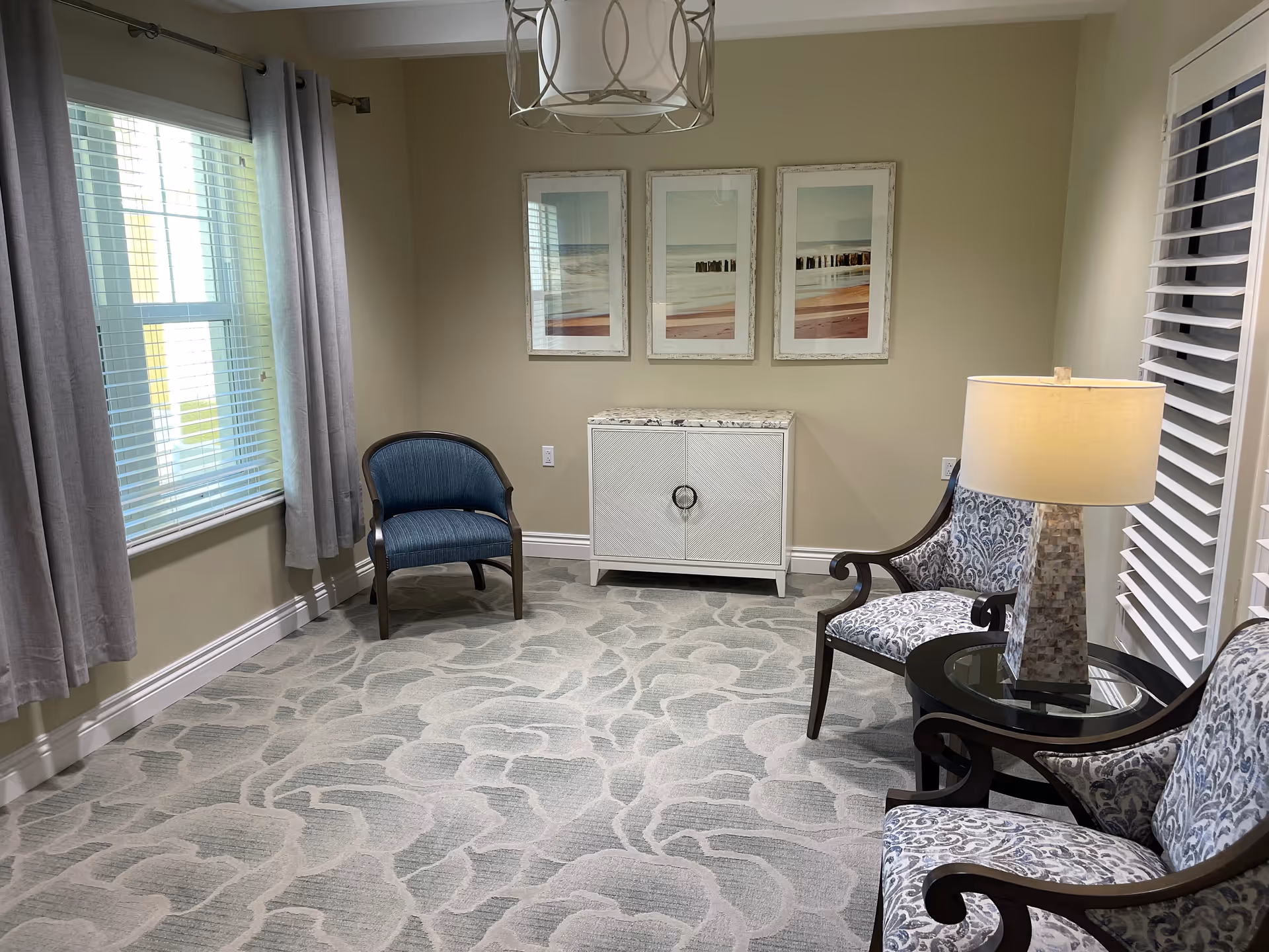 A small sitting area with three chairs, a round side table with a lamp, a white cabinet against the wall, and three framed pictures above the cabinet. The room has beige walls, patterned carpet, two windows with blinds and gray curtains.