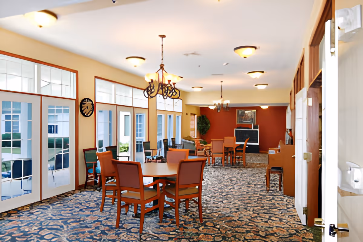 Bright communal dining room with multiple tables and chairs, chandeliers, large windows, and patterned carpet.