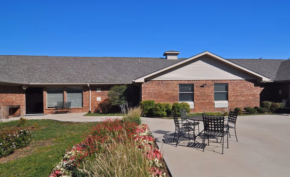 Outdoor patio area at Ridgmar Medical Lodge featuring a concrete seating area with a metal table and four chairs. The patio is surrounded by a garden with various plants and flowers, and the background shows a single-story brick building under a clear blue sky.