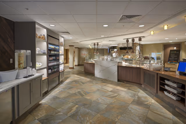 Interior view of a modern dining service area with a tiled floor, a counter with food display, a beverage dispenser, shelves stocked with snacks and drinks, and a staff member behind the counter wearing a white uniform and hat.