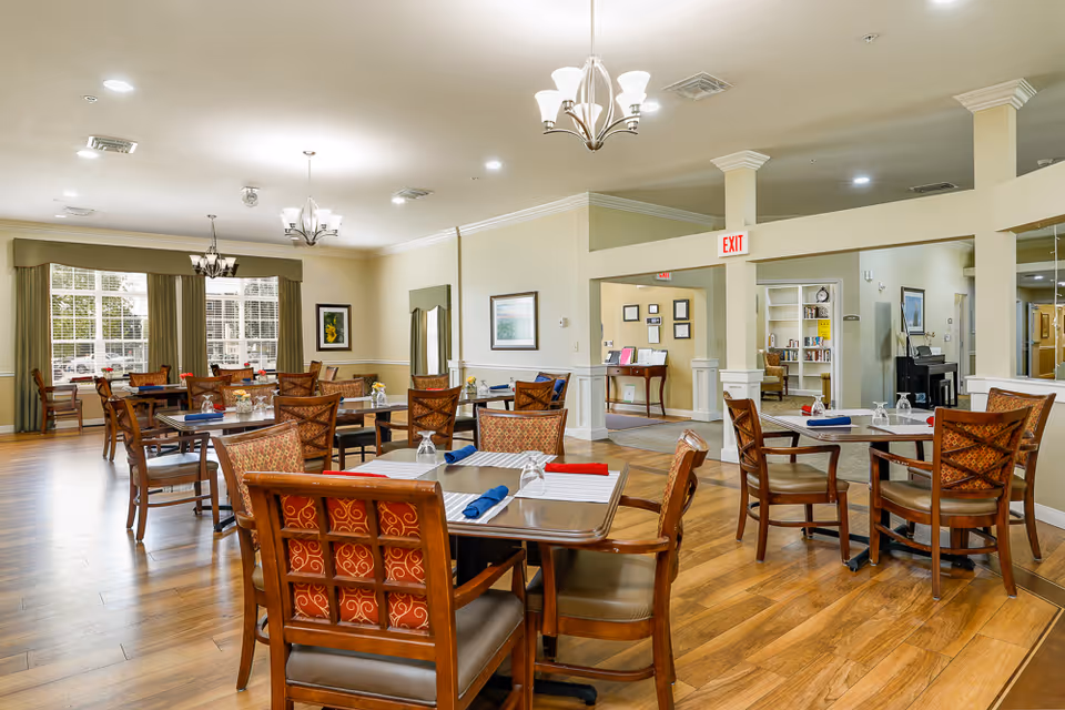 A bright and spacious dining room with multiple wooden tables and chairs arranged neatly. Each table is set with placemats, folded napkins in red and blue, and upside-down glasses. The room features large windows with green curtains, wooden flooring, and several ceiling light fixtures. The walls are decorated with framed artwork, and there is an exit sign above a doorway leading to another room with bookshelves and a piano.